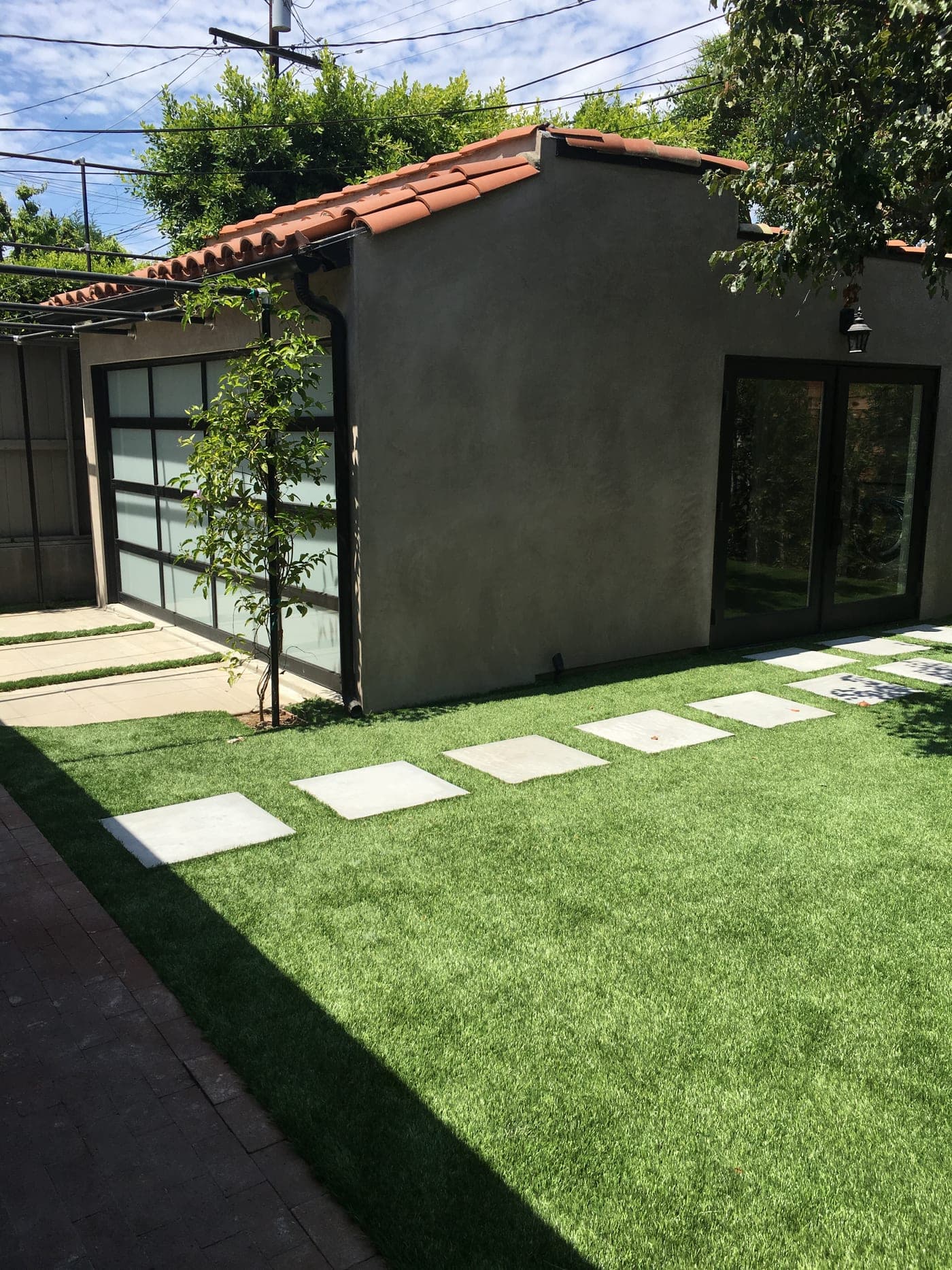 The rebuilt West Hollywood garage from the lawn: smooth stucco, a clay-tile roof, the frosted-glass garage door and new glass doors, with pavers set in the turf