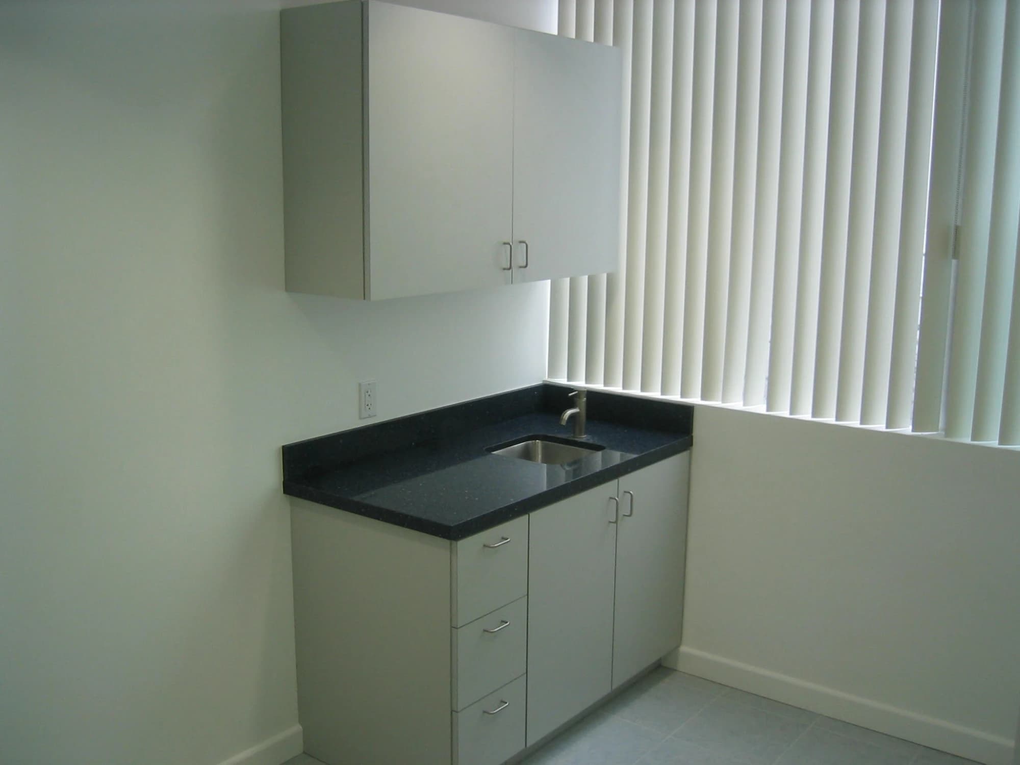 A finished medical exam room with a sink cabinet, dark solid-surface counter and upper cabinets beside a window with vertical blinds