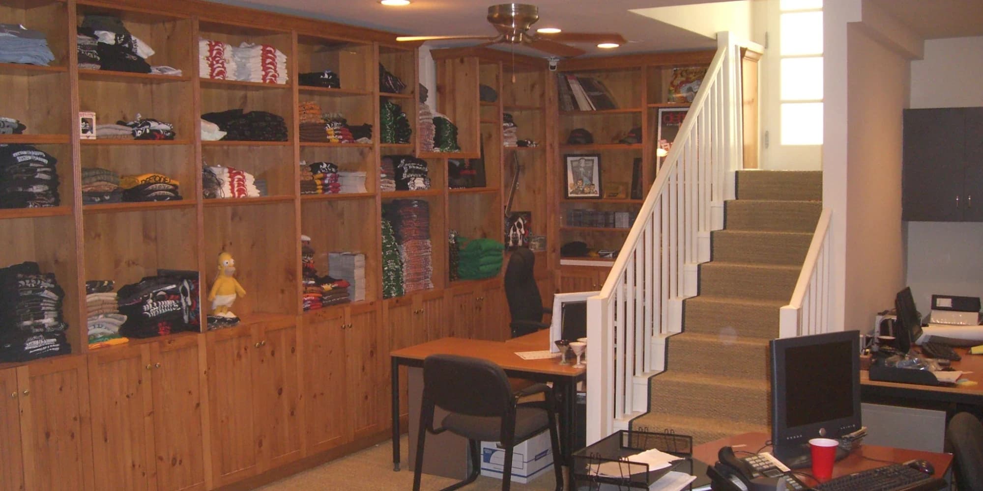 Built-in pine shelving and cabinets lining a merchandise stockroom and office, with folded apparel on the shelves, a staircase and work desks