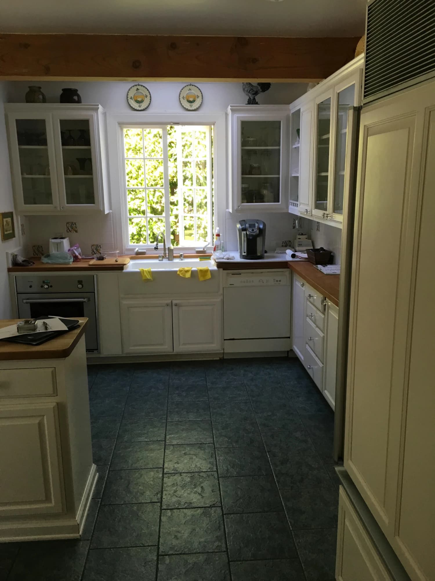 An existing kitchen before its renovation: white cabinetry with glass-front uppers, wood countertops, a farmhouse sink and a dark tile floor