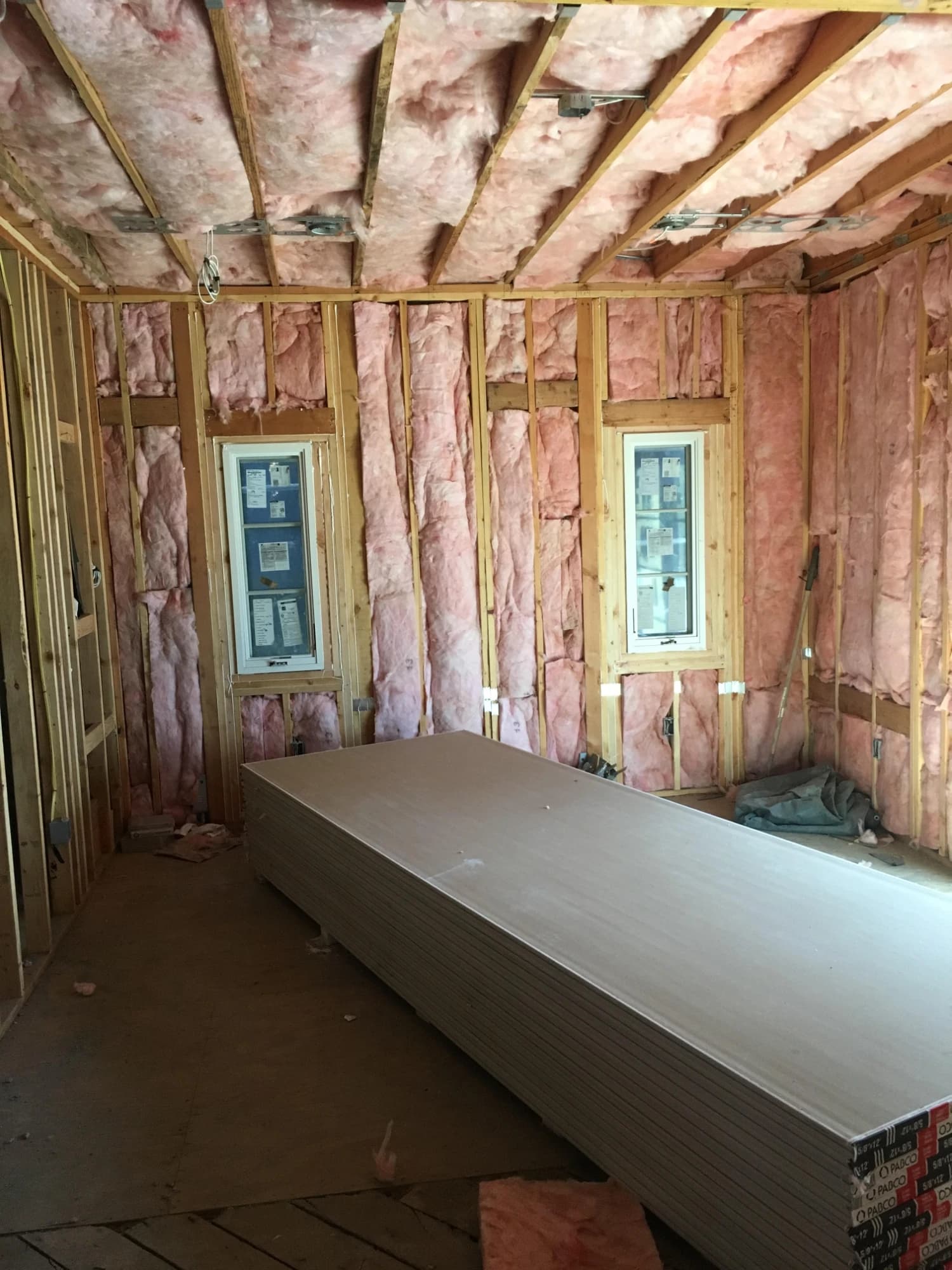 Insulation in the walls and ceiling of a framed room with new windows, and drywall stacked on the floor ready to close the walls