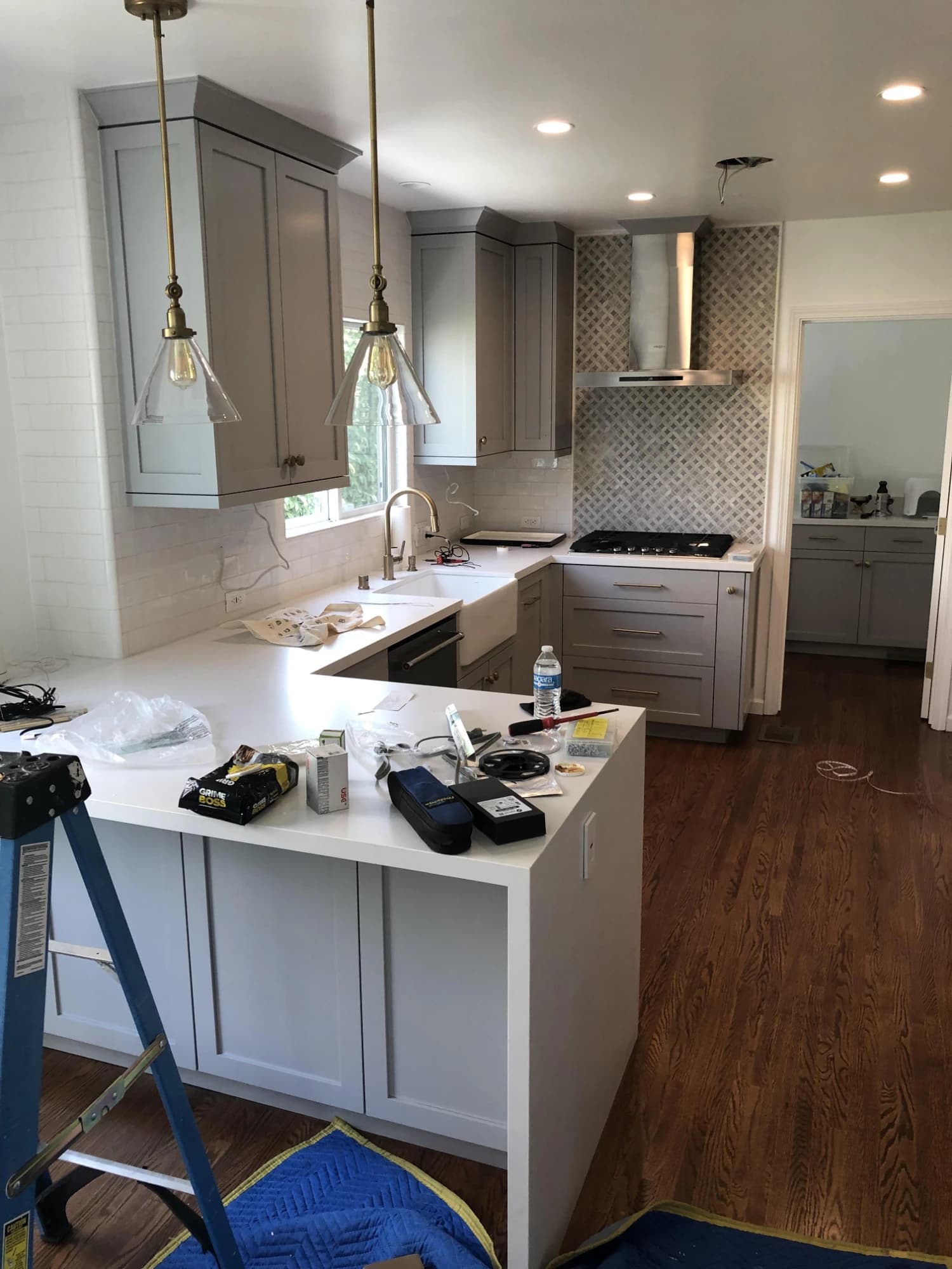 A kitchen nearing completion: gray shaker cabinetry, white quartz counters with a waterfall edge, a farmhouse sink, a patterned tile backsplash, brass pendants and hardwood floors